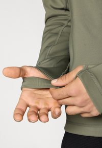 Λαδί μακρυμάνικο πουκάμισο με εφαρμοστά μανσέτες. Ομαλή υφή και επίπεδες ραφές. Το χέρι τραβά τη μανσέτα για ρύθμιση. Το χέρι δείχνει το ύφασμα.