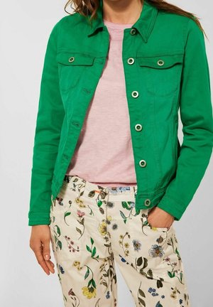 Kobieta ubrana w zieloną zapinaną na guziki kurtkę, jasnoróżową koszulkę oraz kremowe spodnie z kolorowymi kwiatowymi wzorami, jedna ręka w kieszeni.