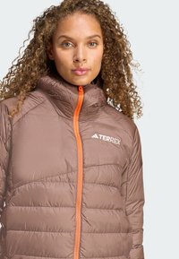 Smeđi izolirani kaput s visokim ovratnikom, narančastim patentnim zatvaračem i quiltiranim uzorkom. Sadrži logotip "TERREX" na lijevoj strani prsnog područja.