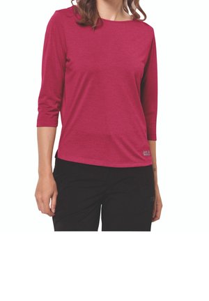 Vrouw draagt een donkerroze shirt met driekwart mouwen en een zwarte broek, staand met ontspannen armen langs haar lichaam tegen een witte achtergrond.