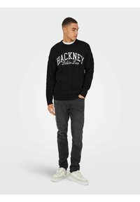 Schwarzer Kabelstrick-Pullover mit Grafik "HACKNEY Dalston Lane", kombiniert mit dunkelgrauen Jeans und weißen Sneakern. Schlichtes Design und bequeme Passform.