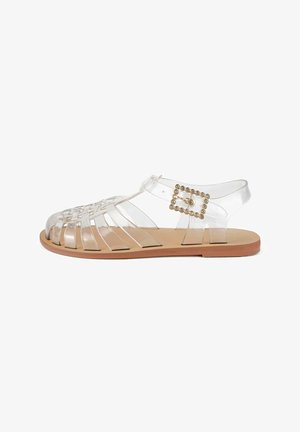 Gennemsigtig PVC sandal med åben tå, flettet design og en guldaccentueret spænde. Flad sål med lys tan-finish.