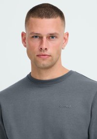Jonge man met kort haar en blauwe ogen, gekleed in een grijze hoodie met het logo "blend", die naar de camera kijkt tegen een lichte, effen achtergrond.