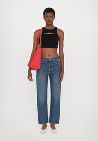 Top noir court avec une découpe, jean droit en denim bleu, chaussures blanches à bout pointu et un sac fourre-tout rouge. Arrière-plan uni.