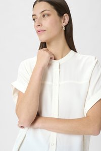 Krémová blůza s krátkým rukávem a mandarínským límcem, která má zapínání na knoflíky vpředu a hladkou, lehkou tkaninu. Jemný horizontální šev.