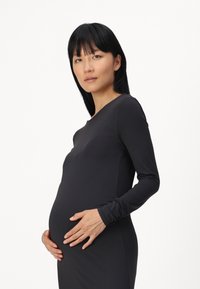 Crna haljina dugih rukava koja je prilagođena tijelu na području torza, s okruglim dekolteom. Model nježno drži vidljivi trudnički trbuh. Glatka tekstura, minimalistički dizajn.
