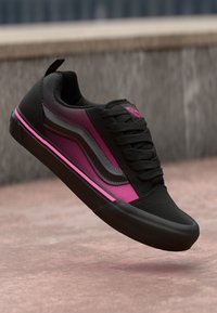 Svarte og rosa lave sneakers med tykke snørebånd og en b�ølgete sidestripe, vist i luften over en betongoverflate.