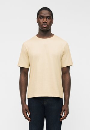 Homme debout devant un fond uni, portant un t-shirt beige à manches courtes et un jean bleu foncé, faisant face à l'avant avec une posture détendue.