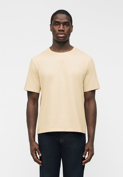 Hombre de pie contra un fondo liso, vistiendo una camiseta beige de manga corta y vaqueros azul oscuro, de frente con postura relajada.