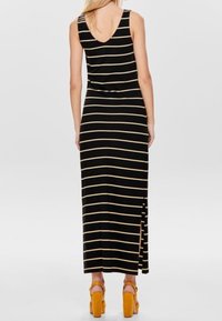 Abito maxi a strisce nere e gialle con scollatura a scoop, spacco laterale e silhouette aderente, abbinato a tacchi alti gialli.