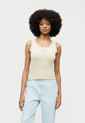 Młoda kobieta o kręconych włosach, ubrana w beżowy bezrękawnik i jasnoniebieskie dżinsy, stoi na tle jednolitego białego tła.