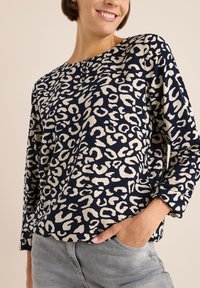 Maglietta a maniche lunghe blu navy con motivo leopardato beige, scollo rotondo e tessuto morbido. Abbinata a pantaloni grigi chiari.