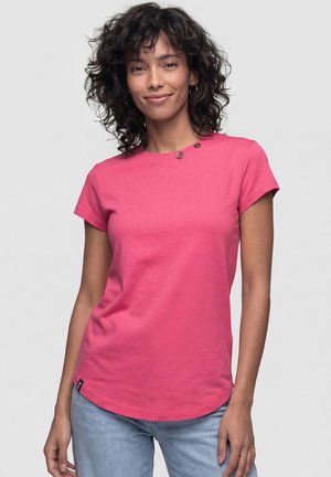 Frau mit lockigem dunklem Haar, die Creolen trägt, ein rosa kurzärmeliges Hemd mit Knöpfen nahe am Kragen und hellblaue Jeans, steht entspannt.