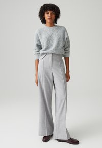 Lichtgrijze, gestructureerde trui met ribbels aan de mouwen; hoogstaande, wijde grijze kraagbroek met ruiten; donkere schoenen maken de outfit compleet.