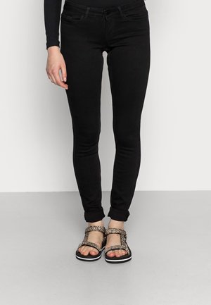 Jambes portant un jean skinny noir et des sandales à bouts ouverts avec des lanières à imprimé léopard, debout sur un sol gris contre un mur blanc.