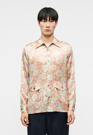 Jonge man draagt een glanzend bloemenoverhemd met lange mouwen in pastel oranje, roze en groen, gecombineerd met donkerblauwe broek met krijtstreep.