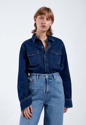 Dunkelblauses Jeanshemd mit zwei Brusttaschen, Knopfverschluss und langen Ärmeln. Kombiniert mit hellblauen, hoch taillierten Jeans.