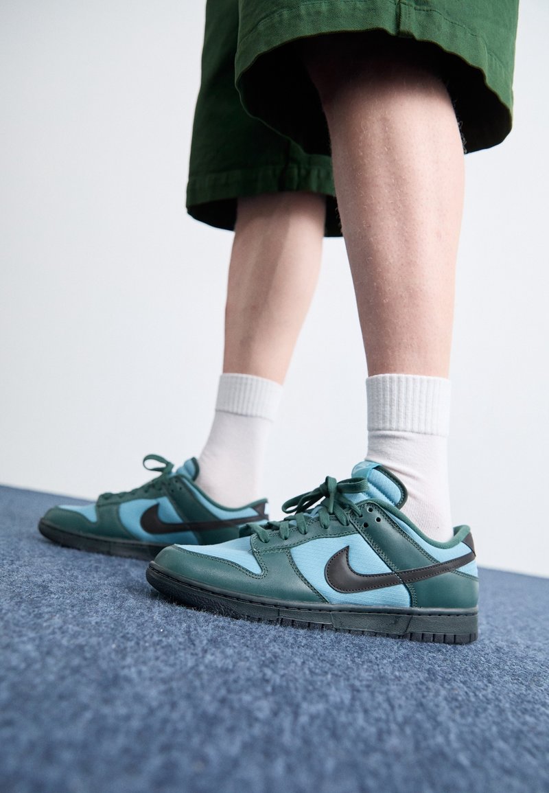 Sneakers Nike Dunk Low v teplé modré a tmavě zelené barvě, s černým swooshem, vyrobené z textilního síťkového a koženého materiálu, a plochou gumovou podrážkou.