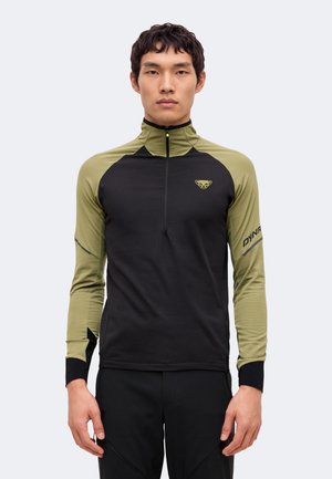 Un homme porte un haut de sport noir et vert olive à manches longues, avec une fermeture éclair courte, sur un fond clair uni.