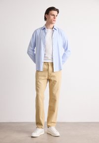 Cămașă albastră deschis, călcată, peste un tricou alb, asortată cu pantaloni bej și adidași albi. Design minimalist, material neted și croială lejeră.
