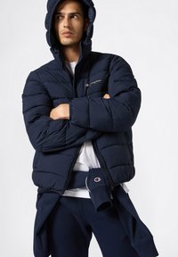 Granatowa kurtka puchowa z kapturem, o pikowanej fakturze i zapięciu na zamek. Logo na piersi. Noszona na białą koszulkę i pasujące spodnie.
