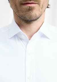 Witte blouse van een textuurstof, met een knoopsluiting en een gestructureerde kraag; close-up weergave die de details van de blouse benadrukt.