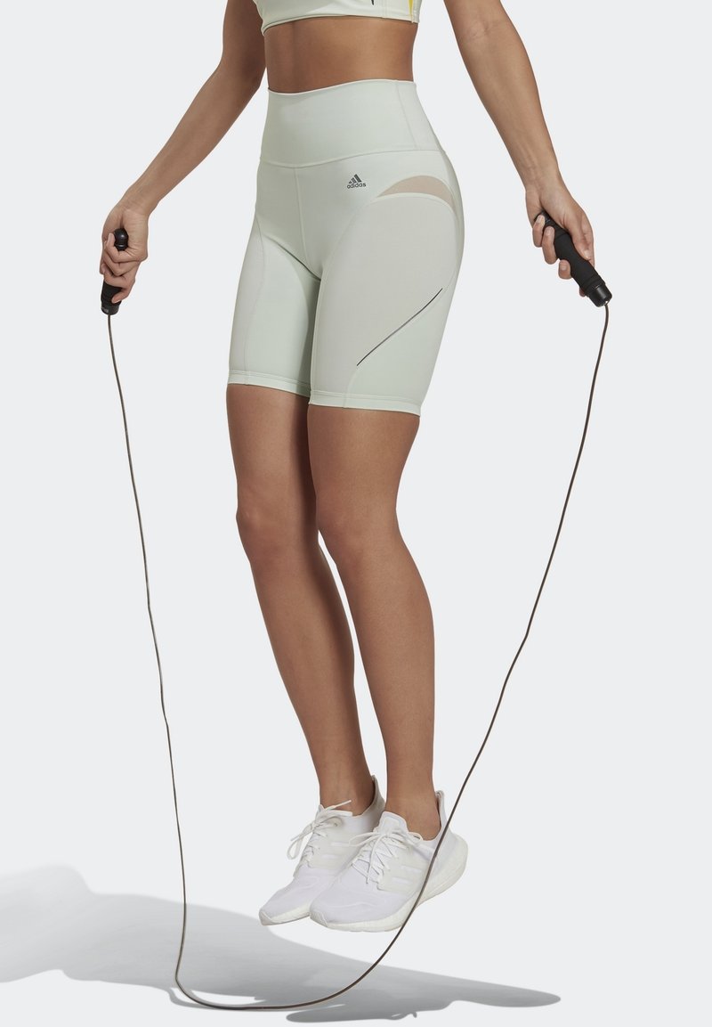 Pantalones cortos deportivos verde claro con cintura alta, panel de malla en contraste y líneas de acento negro. Persona sostiene una cuerda para saltar negra, llevando zapatos blancos.