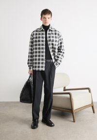 Karirana siva i crna flanel košulja preko crnog pulovera s visokim ovratnikom, uparena s crnim hlačama i elegantnim crnim cipelama. Drži crnu torbu.