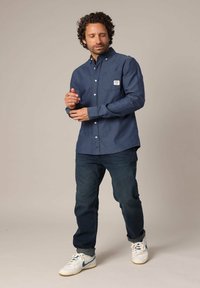 Overhemd met knoopsluiting in donkerblauwe stof met lange mouwen en een borstzak; gecombineerd met donkere spijkerbroek en witte sneakers met blauwe accenten.