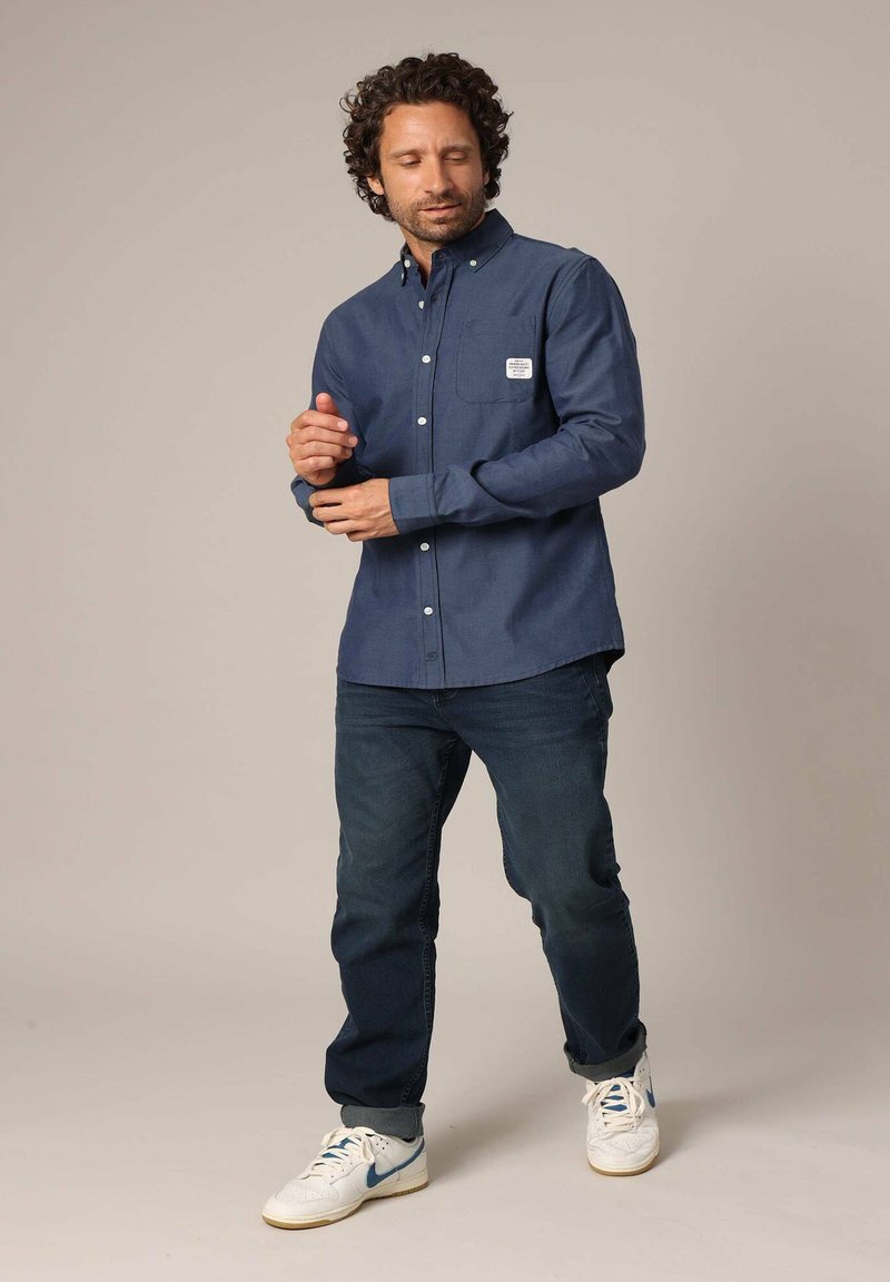 Overhemd met knoopsluiting in donkerblauwe stof met lange mouwen en een borstzak; gecombineerd met donkere spijkerbroek en witte sneakers met blauwe accenten.