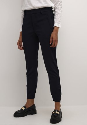 Pantalons ajustés navy en tissu lisse, avec poches latérales et zips aux chevilles. Portés avec des mocassins noirs.
