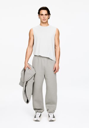 Ung mand stående iført lys grå ærmeløs skjorte og matchende joggingbukser, holdende en foldet grå hættetrøje i den ene hånd.