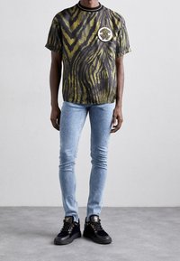 Camiseta a rayas negra y verde oliva con un logo redondo, combinada con unos jeans skinny azul claro y zapatillas negras con detalles en dorado.