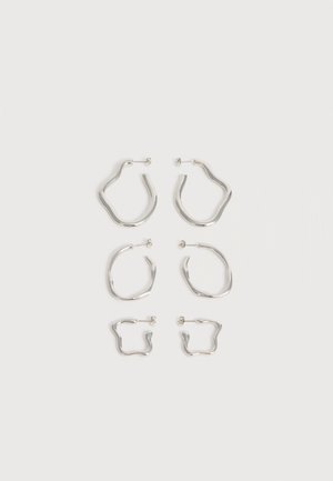 Trois paires de boucles d'oreilles en argent de forme irrégulière, en grandes, moyennes et petites tailles, disposées verticalement sur un fond blanc.