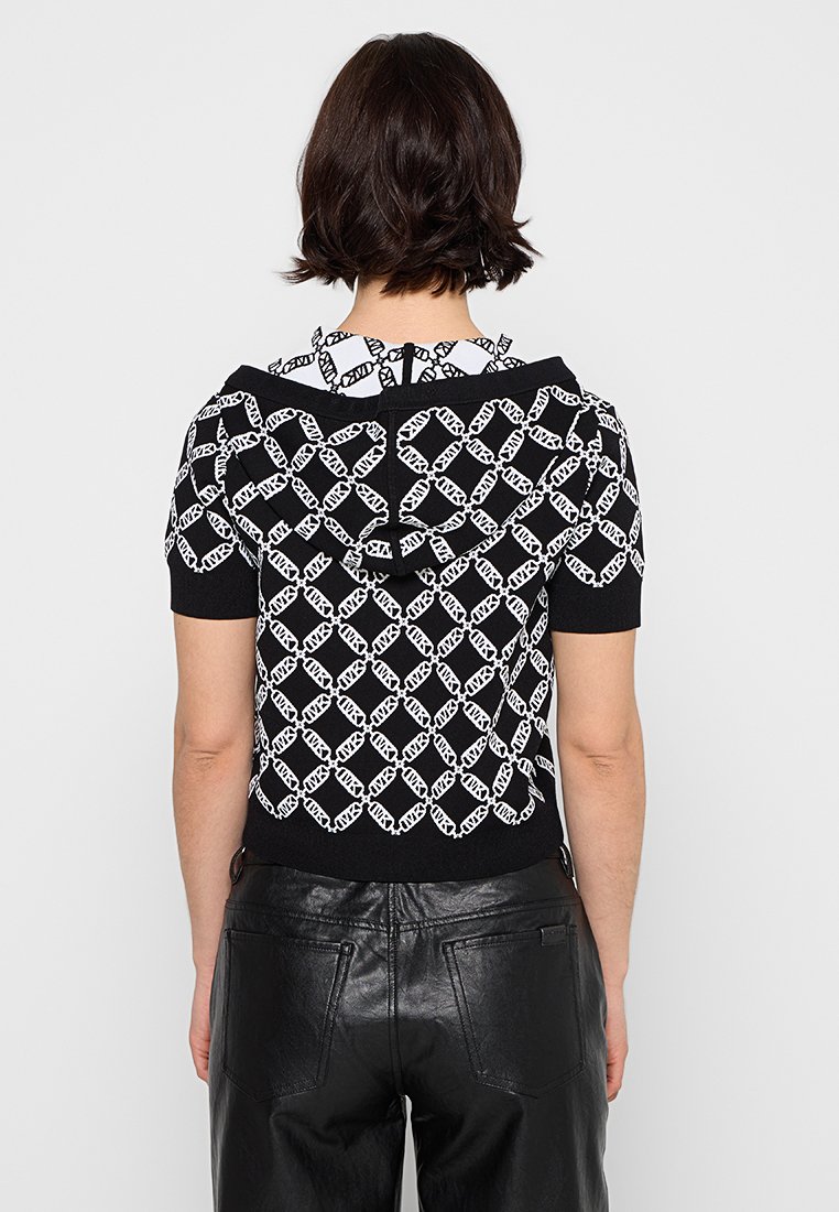 Zwart gebreid top met een wit kettingpatroon, korte mouwen en een capuchon. Bevat een striksluiting aan de achterkant en een geribbelde zoom. Gecombineerd met zwarte faux-lederen broek.