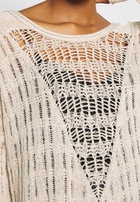 Pull en maille beige à tricot lâche, avec un col large et une décolleté en V prononcé, porté sur un haut noir, mettant en valeur les motifs texturés.