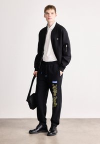 Μαύρο μπουφάν bomber με φερμουάρ, πάνω από λευκό πόλο. Μαύρα φόρμα με μπλε λεπτομέρειες και κίτρινο κείμενο. Μαύρα παπούτσια και τσάντα ώμου.