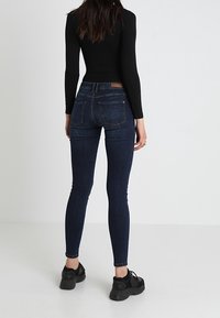 Kvinna iklädd mörkblå slim fit jeans, svart långärmad topp och svarta sneakers, stående vänd bort mot en enkel vit bakgrund.