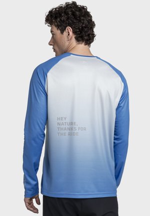 Mann mit lockigem Haar, der ein blau-weißes Langarmshirt trägt, auf dessen Rücken der Text "HEY NATURE, THANKS FOR THE RIDE" steht.