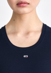 Marinblå ribbstickad tröja med rund halsringning och en liten broderad logotyp med röda och vita detaljer på vänster bröstområde.