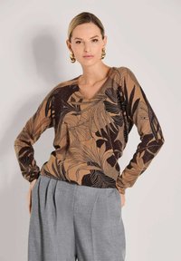 Vrouw in een bruine gebloemde langeklassige top met zwarte bladdessins, grijze geplooide broek en grote gouden hoepeloorbellen, poseert met handen op de heupen.