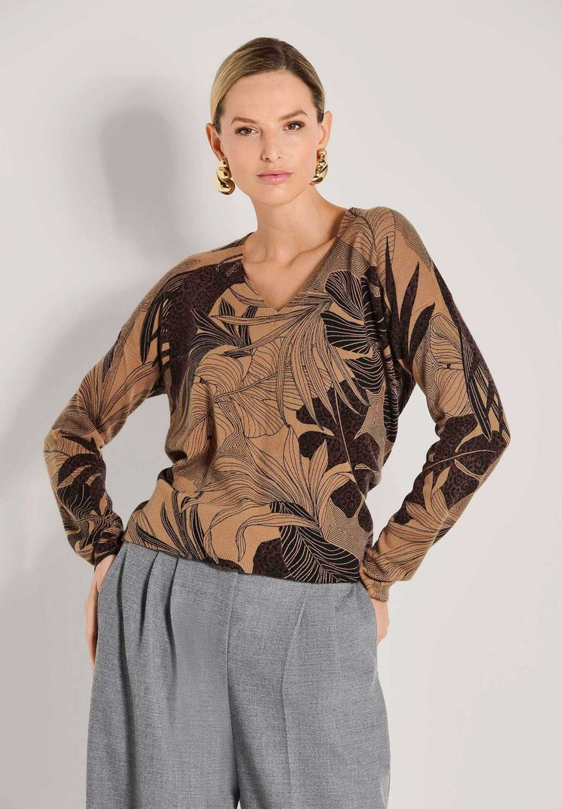 Vrouw in een bruine gebloemde langeklassige top met zwarte bladdessins, grijze geplooide broek en grote gouden hoepeloorbellen, poseert met handen op de heupen.