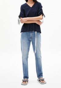 Persona con indosso una maglietta blu navy a V, jeans chiari con cordoncino bianco e infradito grigie imbottite, con le braccia incrociate.