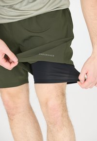 Groene atletische shorts over zwarte compressieshorts, voorzien van een logo op de buitenste shorts, soepel materiaal en een ontspannen pasvorm.