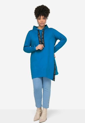 Tunique à capuche bleue en tissu doux, dotée d'un cordon noir avec du texte et d'un ourlet asymétrique, associée à un jean en denim clair et des bottines beige.