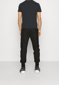 Pantalons cargo noirs avec poches latérales, associés à un t-shirt sombre et des chaussures de randonnée grises, vus de l'arrière sur un fond uni.