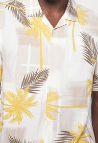 Gros plan d'une personne portant une chemise à manches courtes boutonnée avec des motifs de palmiers et de feuilles jaunes et bruns sur un fond clair.