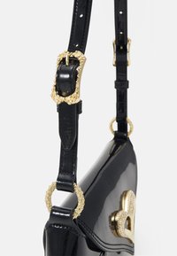 Svart lackläderhandväska med en glänsande yta, med guldfärgade detaljer och en texturerad spänne på den justerbara axelremmen.