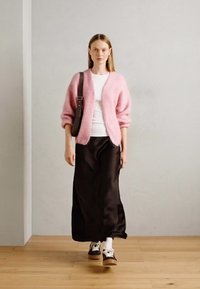 Cardigan fluffy rosa su una maglietta bianca, abbinato a una gonna maxi in satin nera e scarpe da ginnastica marroni con calze bianche. Visibile una semplice borsa a spalla.