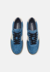Blå ruskinnsneakers med en kremfarget sidestrek, svarte snøringer og gummisåle. Merkenavnet "SAMSOE" synlig på innersålen.